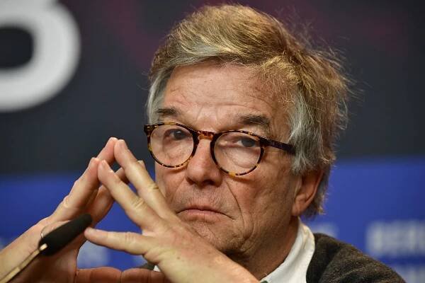 A man wearing round tortoiseshell glasses tents his fingers in front of his face.