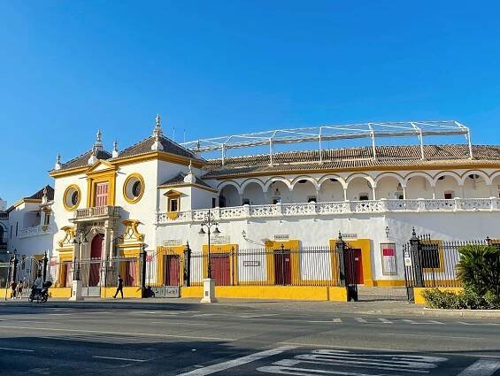 Plaza de Toros - A large, ornate building with a yellow and white facade under a clear blue sky. The structure features arches, circular windows, and a detailed roof with decorative elements as people stroll by on the bustling street.