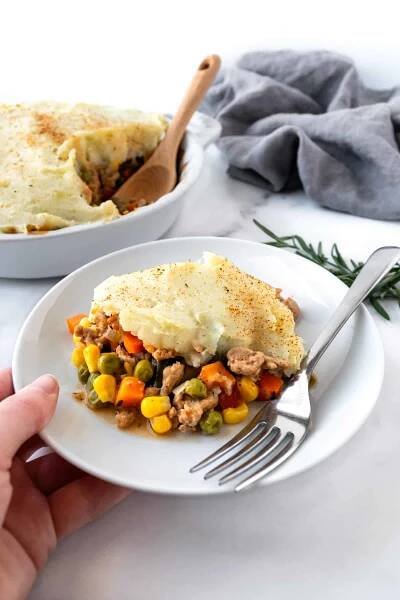A hand holding a white plate with a piece of shepherds pie on it with a fork.