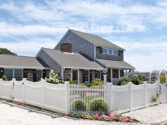 a short white spaced picket fence in front of a shingled beach home with crushed shells in the foreground