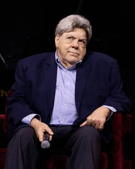 George Wendt sits in a chair holding a microphone
