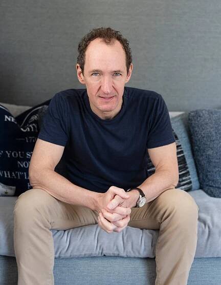 A man in a blue T-shirt and khaki pants looks into the camera while sitting on a couch.