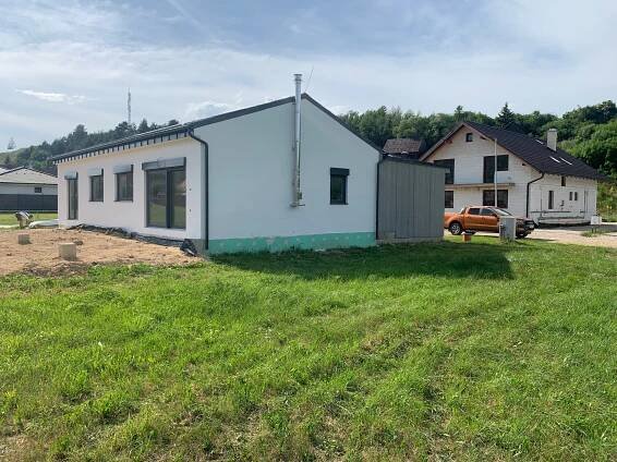 Zadný pohľad na montovaný drevodom v obci Vikartovce s bielou fasádou, antracitovými oknami, sedlovou stechou a parciálnymi dreveným obkladom v prístrešku s navezenou a vyrovnanou zeminou na pozemku