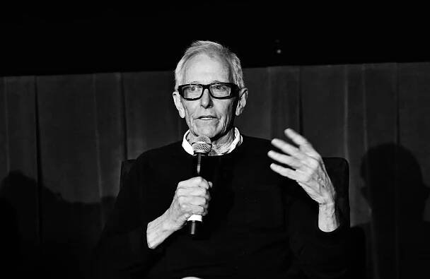 A black-and-white photo of Jim Abrahams, with gray hair and glasses, holding a microphone with one hand and gesturing with the other.