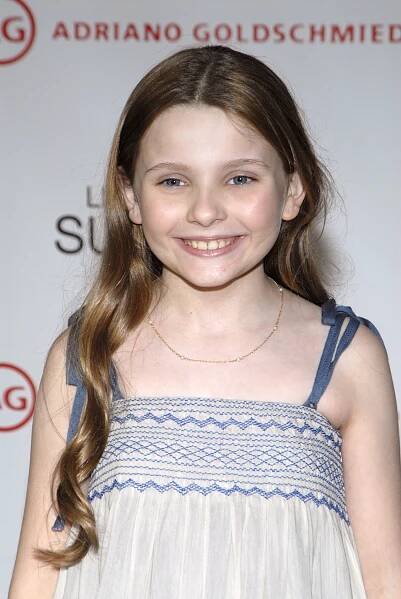 A person smiles while standing on a red carpet, wearing a sleeveless dress with a detailed upper pattern and long hair