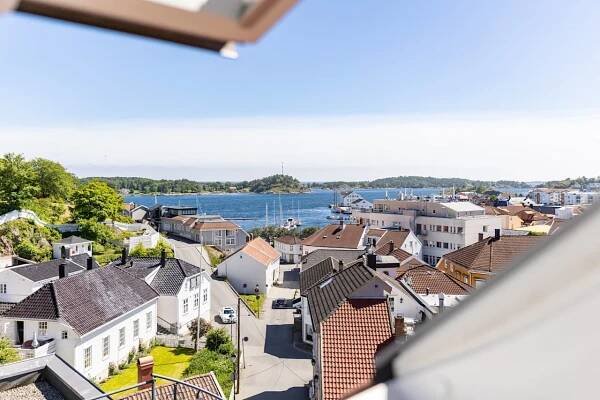 A coastal town with white houses, red roofs, a street, and a car, framed by an open window, overlooking a bay under a clear sky.