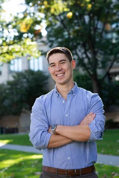 A man in a blue shirt stands outside in front of a building on the Harvard Law School campus.