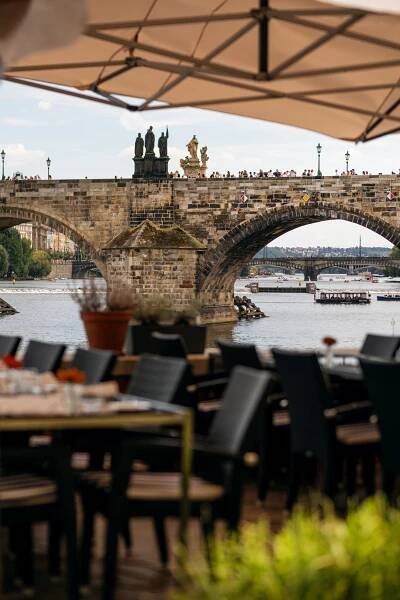 Pohled na historický kamenný most přes řeku z venkovní restaurace se stoly a židlemi v popředí. Na vrcholu mostu jsou vidět sochy a na vodě několik lodí. Scéna se odehrává za jasného, částečně zamračeného dne.