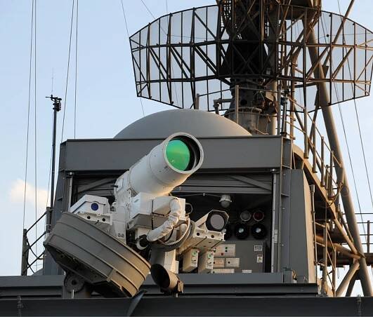 A naval laser weapon system mounted on a military ship, featuring a prominent white laser cannon with a green-tinted optical lens, set against a backdrop of radar equipment, antennas, and support structures.