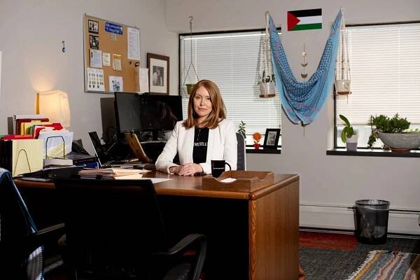 A woman sits at her desk in an office decorated with plants and a Palestinian flag on the wall.