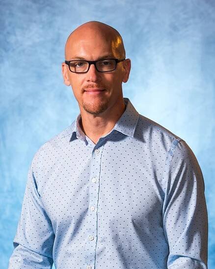 A bald man wearing glasses and a blue shirt is standing in front of a blue background.