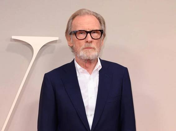 bill nighy standing on a red carpet, wearing a black suit and white dress shirt with no tie. he has on black glasses and a neutral expression on his face