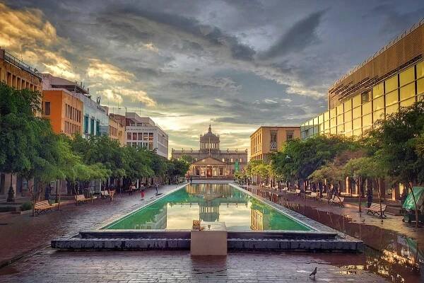 In the center, a long pool of water flanked by modern buildings, some colorful, and some with glass windows, all leading to a neoclassical looking building with a large dome in the distance beneath a cloudy sky in Guadalajara, Mexico. 