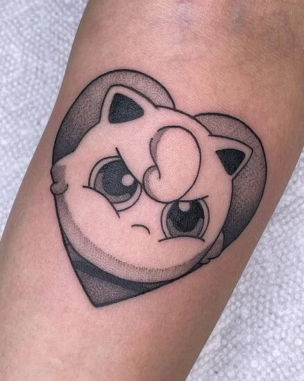 A black and grey ink tattoo of Jigglypuff in a heart shape on someone's forearm. Jigglypuff has an annoyed expression.