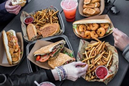 Hands reach for fries across a table that’s covered in black carryout containers filled with fries, pressed sandwiches, and shrimp sandwiches. 