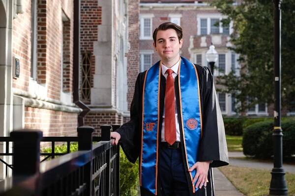 A graduate wearing a orange tie, blue stole and black gown leans against a black fence with a brick building in the background.