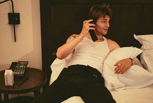 My Life With the Walter Boys actor Noah LaLonde laying on his hotel room bed talking on the phone. He is wearing a white tank top and black pants by Rhude.