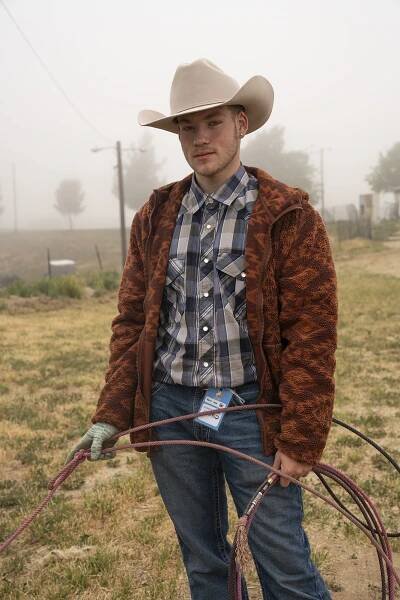 Portrait of a person in tan cowboy hat, blue plaid shirt, and brown fleece jacket, holding a lasso.
