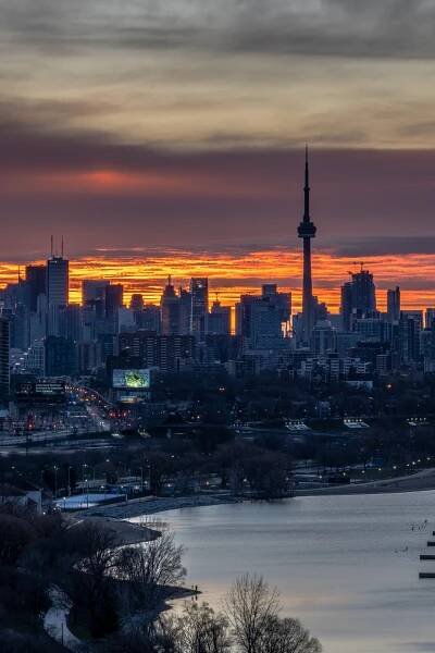 A view over a city skyline with a body of water to the right. A spire towers above the skyscrapers. The sky is turning a bright orange.