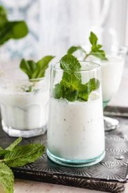 Several clear glasses filled with Ayran, a yogurt and mint drink, placed on a silver tray and topped with fresh mint.