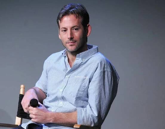 Person sitting on a director's chair, casually dressed in a striped button-up shirt, holding a microphone