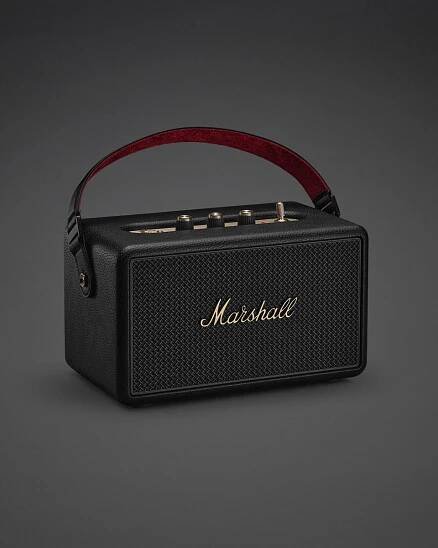 The Marshall Kilburn III Black and Brass portable speaker, featuring a red handle, gold control knobs, and the iconic logo on the front grille, is displayed against a dark background.