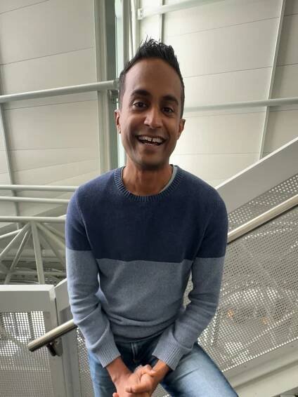 Jeremy Fernandez stands in a stairwell in a blue jumper and smiles at the camera.