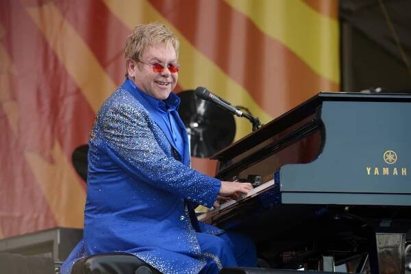 Elton John během živého vystoupení, sedí u černého koncertního klavíru.