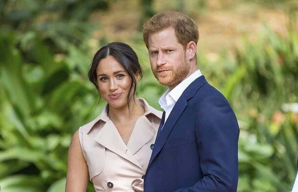 Harry and Meghan standing outside, she smiles, he scowls. 