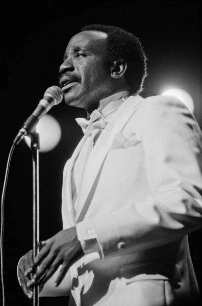 A black-and-white photo of Jerry Butler, a man with close-trimmed hair and a mustache, standing at a microphone and singing. He is wearing a white tuxedo.
