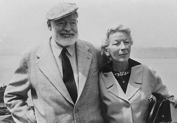 A black and white photograph shows an aged Ernest Hemingway wearing a light-colored plaid suit and a plaid cap. To his right is his wife, Mary, also wearing a light-colored suit, with a dark blouse. She is holding a purse.
