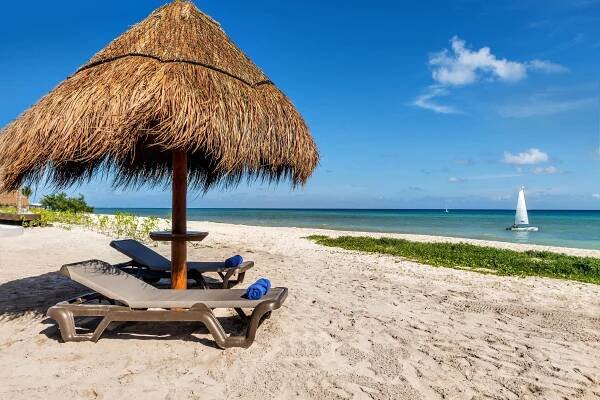 Ocean Riviera Paradise All Inclusive Playa del Carmen Exterior photo The photo shows a beautiful tropical beach scene on a sunny day. The foreground features a thatched-roof beach umbrella providing shade over two lounge chairs. The chairs are made of a light-colored material and have blue towels draped over the armre