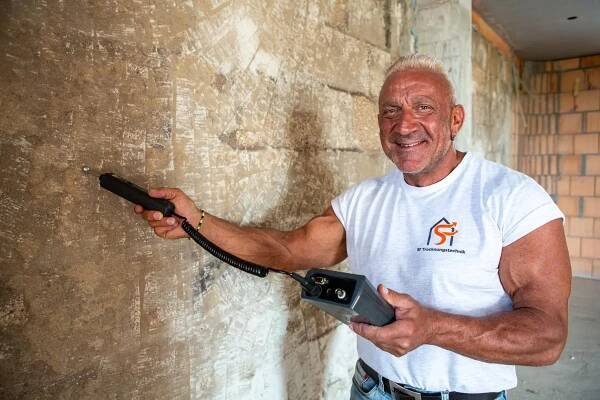 Porträtbild von Stefan Friedrich, Geschäftsführer SF Trocknungstechnik, bei der Arbeit. Er hält ein Messgerät an eine Wand und lächelt in die Kamera.