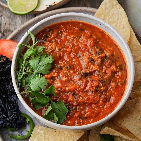 A bowl of red salsa rojo garnished with cilantro, surrounded by tortilla chips, a slice of tomato, lime wedge, and dried pepper on a wooden surface.