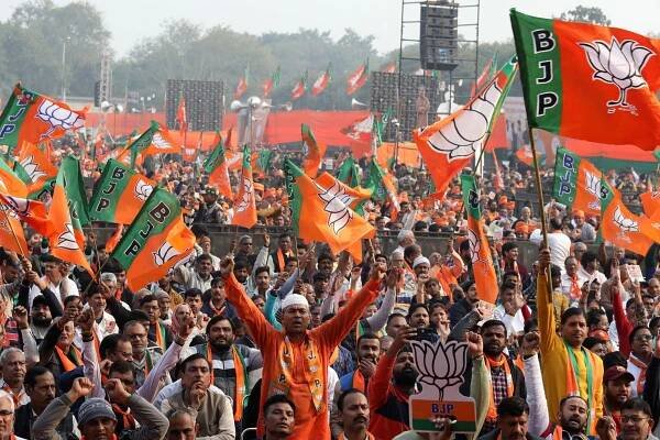 A large crowd of men, many waving their arms or holding up green and saffron flags that say "BJP."
