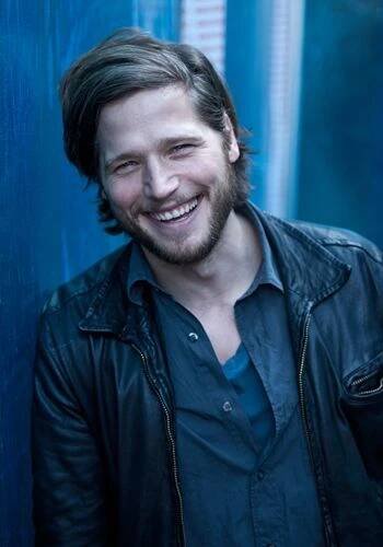 a smiling man in a black leather jacket leaning against a blue wall with his hand on his hip