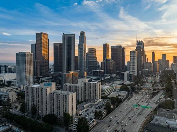 A view of the downtown Los Angeles skyline at sunset, with tall skyscrapers, city buildings, and a busy freeway in the foreground under a partly cloudy sky.