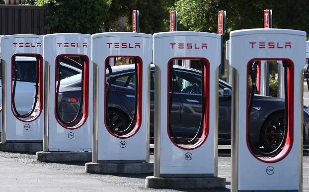A dark blue Tesla vehicle sits parked at a Tesla Supercharger station, behind a row of silver and red chargers.