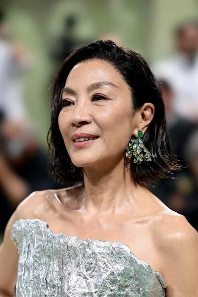 Michelle Yeoh in a silver gown, green statement earrings, and her hair in a wet hair flip style
