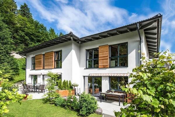 Moderne zweistöckige Fertighaus mit weißer Fassade und Holzfensterläden, umgeben von grünen Bäumen und gepflegtem Garten, in einer sonnigen Landschaft in Österreich. Perfekt für Bauprojektplanung bei WOLF Haus.