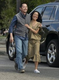 Steven Yeun and Ali Wong walk together on the set of BEEF. Yeun wears jeans and a grey vest and Wong wears a khaki matching set. 