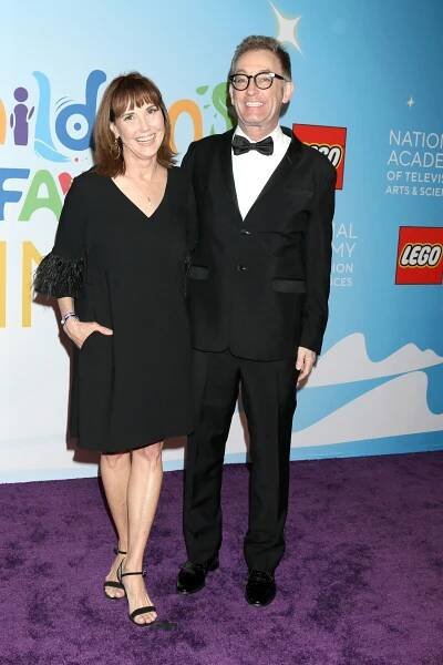 Two people in formal attire at a media event. The woman is in a black dress, and the man is in a tuxedo