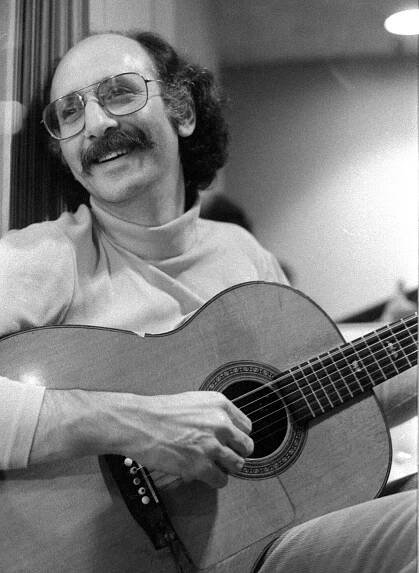 A black-and-white photo of a balding man with shaggy dark hair on the back of his head, a bushy mustache and glasses smiling and playing an acoustic guitar.