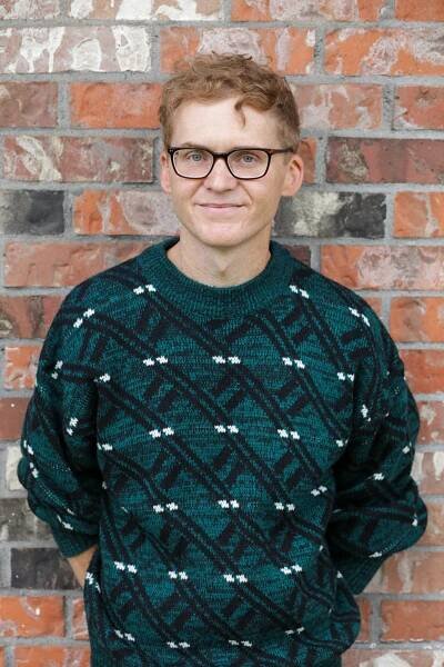 the author stands against a brick wall. He is wearing glasses and a green patterned sweater.