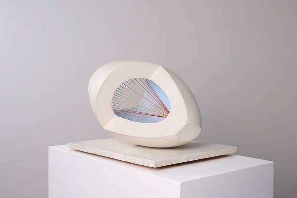 A modern abstract sculpture with a smooth, off-white oval shape featuring a hollow center. Inside, red strings are stretched in a geometric pattern against a light blue background. The sculpture is placed on a square white base.