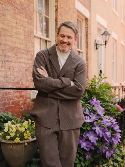 Michael Arden stands with arm crossed, backed by potted flowers in bloom.