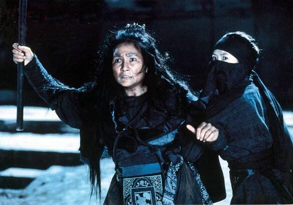 A woman with flowing black hair holds a sword while a woman wearing a mask stands behind her,