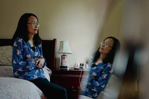 Annie Chen sits on her bed at home. She wears a blue patterned shirt and glasses. Her reflection is in a nearby mirror.