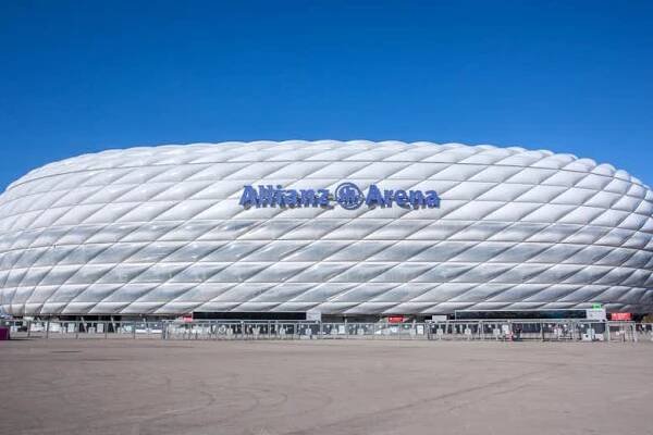 Velký bílý stadion označený „Allianz Arena“ proti jasně modré obloze s rozsáhlým prázdným chodníkem v popředí.