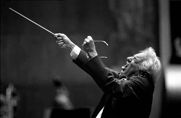 Leonard Bernstein passionately leads an orchestra, raising his baton in one hand and holding glasses in the other.